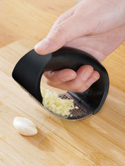 Hand using a garlic press on a wooden cutting board with a whole garlic clove beside it.