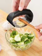 Hand using a garlic press to crush garlic over a bowl of chopped cucumbers on a wooden cutting board.