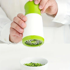 Hand holding a green and white manual herb grinder with a bowl of ground herbs below on a white background