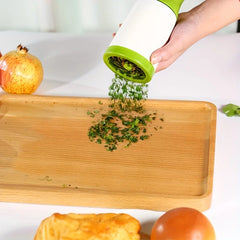 Hand using a green herb grinder to sprinkle herbs onto a wooden cutting board with fruits and vegetables in the background.