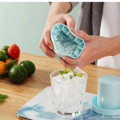 Person using a silicone ice cube tray to add ice cubes to a glass of iced tea on a wooden table with fruits in the background.