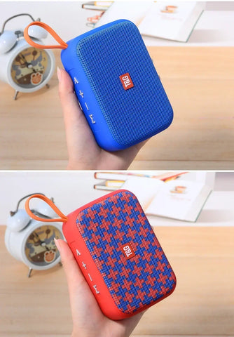 Blue and red portable speaker held by a hand on a wooden surface with a clock in the background.