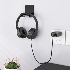 Black headphones hanging on a wall-mounted holder with a cable plugged into an outlet, next to a small potted plant.